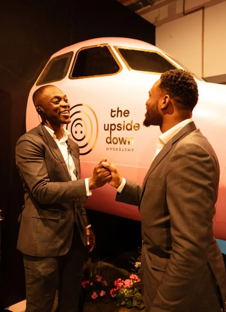 Twee mannen in pak schudden elkaar de hand voor een roze vliegtuigwand met het logo van The Upside Down Amsterdam.