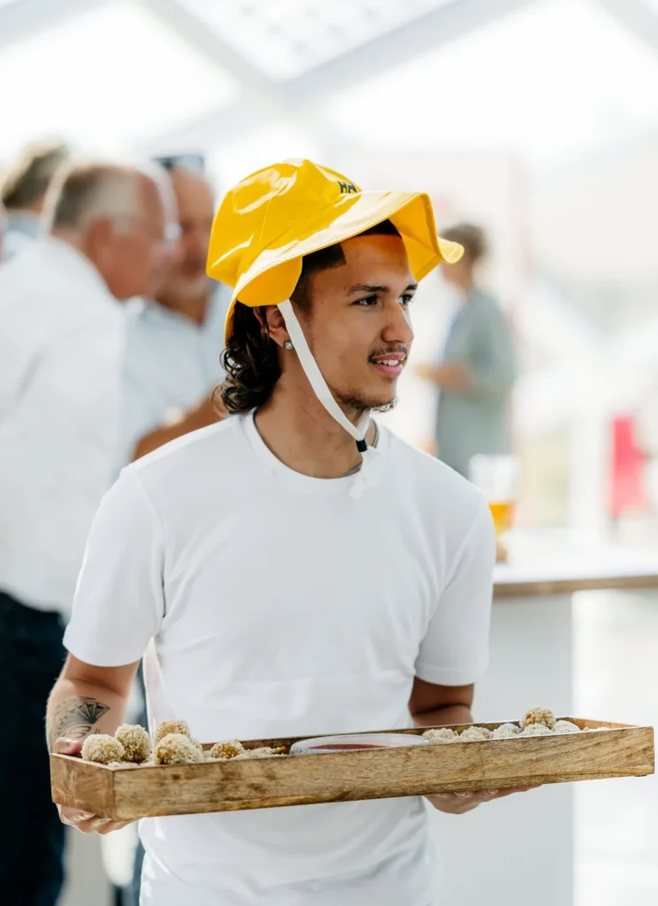 Man met een gele vissershoed serveert hapjes op een houten dienblad tijdens een evenement.